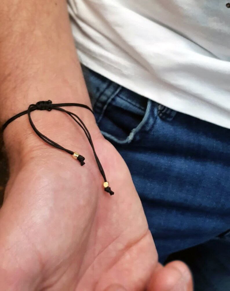 May include: Black adjustable bracelet with gold-colored beads. The bracelet is tied with a sliding knot closure. The bracelet is worn on a wrist, with a white shirt and blue jeans in the background.