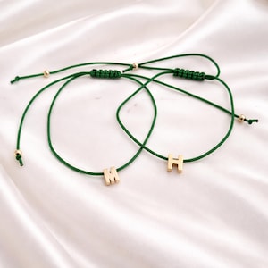 May include: Two adjustable green cord bracelets with gold-colored metal letter charms. One bracelet has the letter "M" and the other has the letter "H". The bracelets have gold-colored beads and macrame knots.