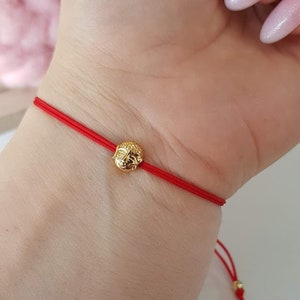 May include: A red string bracelet featuring a small, gold-colored Buddha head charm. The bracelet is tied around a wrist, showcasing the delicate design and vibrant color of the cord and charm.