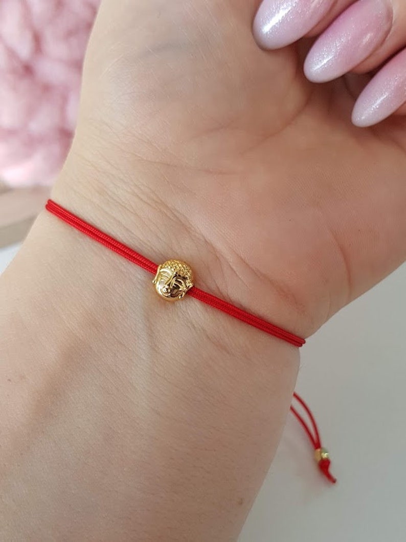 May include: A red string bracelet featuring a small, gold-colored Buddha head charm. The bracelet is tied around a wrist, showcasing the delicate design and vibrant color of the cord and charm.