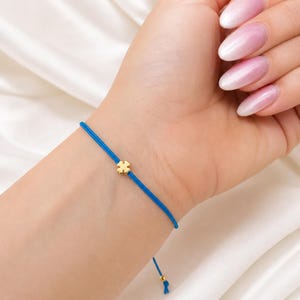 May include: A blue string bracelet with a gold-colored four-leaf clover charm. The bracelet is tied on the wrist. The background is a white, silky fabric.