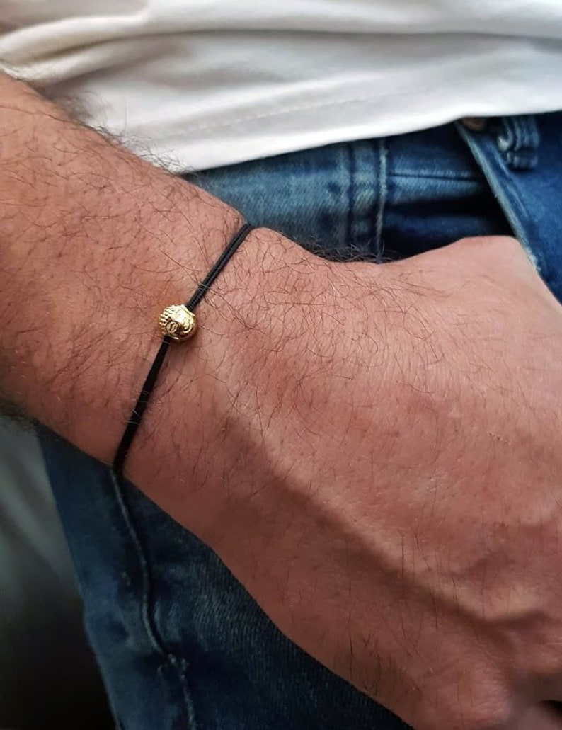May include: A black cord bracelet with a gold-colored bead. The bead has a textured surface and a small design. The bracelet is worn on a wrist against a background of blue denim jeans and a white shirt.