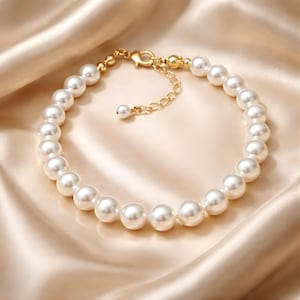 May include: A delicate pearl bracelet with a gold-colored clasp and chain. The bracelet features multiple round, white pearls strung together. The background is a soft, satin-like fabric in a light beige color.