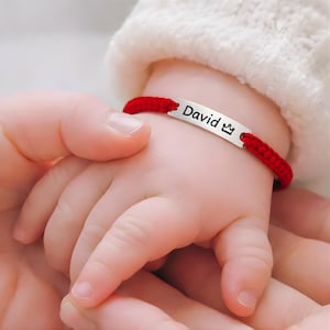 Può includere: Un braccialetto rosso intrecciato con una targhetta argentata con la scritta "David" e un piccolo simbolo di corona. Il braccialetto è al polso di un bambino, tenuto dalla mano di un adulto. Il bambino indossa un maglione bianco.