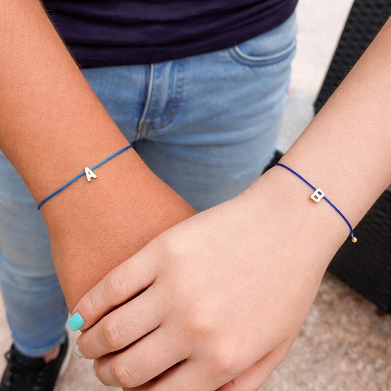 May include: Two blue string bracelets, each with a single silver letter charm. One bracelet features the letter "A", the other the letter "B". The bracelets are worn on wrists, with one person holding the other's hand.