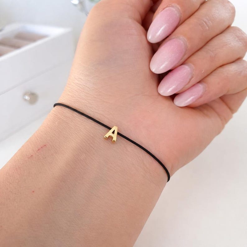May include: A black cord bracelet with a gold-colored letter "A" charm. The bracelet is simple and delicate, suitable for everyday wear. The charm is small and centered on the black cord.