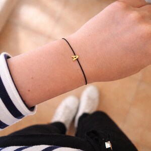May include: A gold-colored letter "A" charm bracelet on a black cord. The bracelet is worn on a wrist, with a striped sleeve and white sneakers visible in the background. The bracelet is a simple, minimalist design.