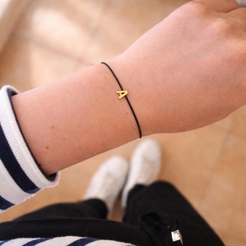 May include: A gold-colored letter "A" charm bracelet on a black cord. The bracelet is worn on a wrist, with a striped sleeve and white sneakers visible in the background. The bracelet is a simple, minimalist design.