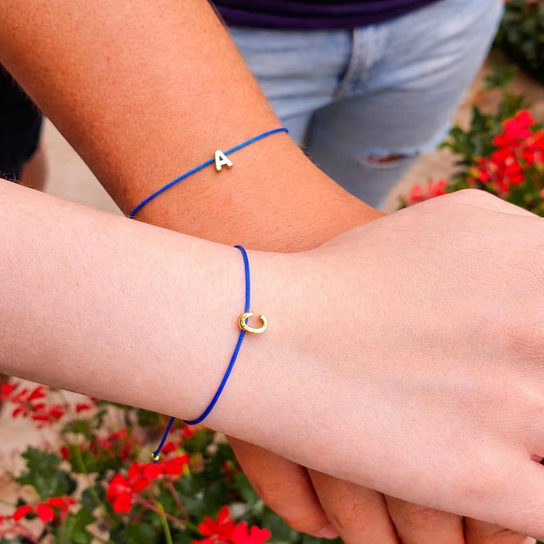 May include: Two blue string bracelets, each with a gold-colored initial charm. One bracelet features the letter "A", and the other has the letter "C". The bracelets are worn on wrists, with a background of red flowers and blue jeans.