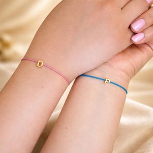 May include: Two wrists each adorned with a delicate bracelet. One bracelet features a pink cord with a gold letter "D", the other a blue cord with a gold letter "P". The bracelets are simple and elegant, perfect for everyday wear.