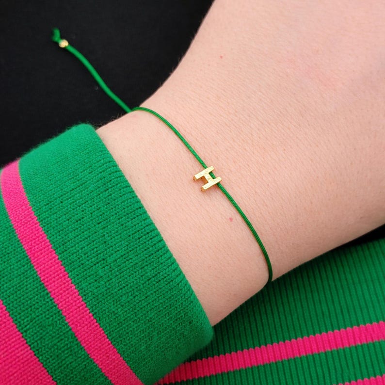 May include: A gold-colored letter "H" charm bracelet on a green cord. The bracelet is worn on a wrist, with a green and pink striped sleeve in the background. The bracelet is simple and elegant.