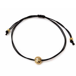 May include: A black cord bracelet featuring a gold-colored Buddha head charm. The adjustable bracelet has gold-colored beads at the ends of the cords. The Buddha head is detailed with facial features.