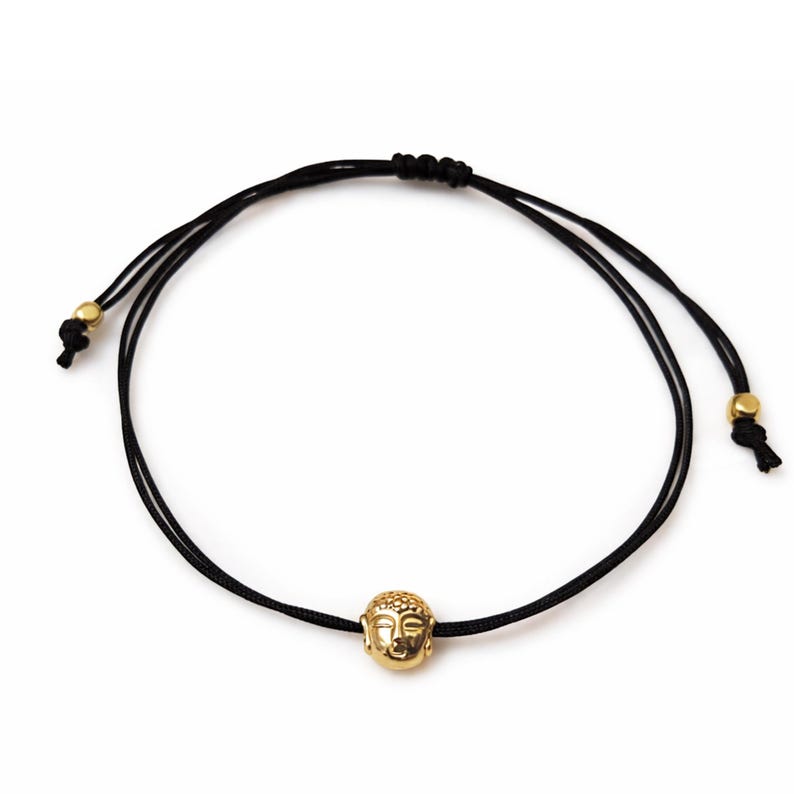 May include: A black cord bracelet featuring a gold-colored Buddha head charm. The adjustable bracelet has gold-colored beads at the ends of the cords. The Buddha head is detailed with facial features.