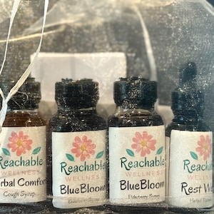 Può includere: Una collezione di quattro piccole bottiglie di vetro con tappi neri, ciascuna etichettata con "Reachable Wellness" e un design floreale. Le bottiglie contengono sciroppi alle erbe, tra cui "Herbal Comfort" e "BlueBloom", presentate in un sacchetto bianco trasparente.