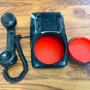 May include: A vintage black telephone handset, base, and dial cover with a bright red interior. The handset has a coiled cord. The phone is set against a wooden background.