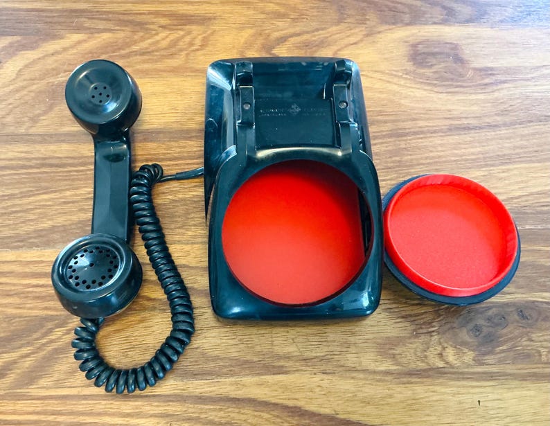May include: A vintage black telephone handset, base, and dial cover with a bright red interior. The handset has a coiled cord. The phone is set against a wooden background.