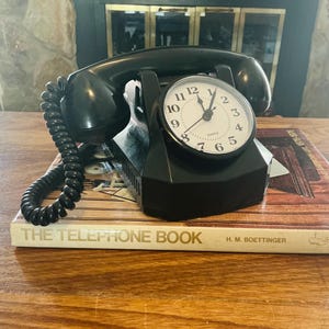 May include: A black desk clock designed to resemble a vintage telephone with a handset resting on top. The clock face is white with black numerals and hands, featuring the word quartz. It sits atop a book titled The Telephone Book by H. M. Boettinger.