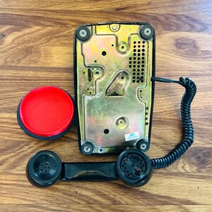 May include: A vintage black telephone with a red button and a coiled cord. The phone's base is gold-colored, and the handset is black. The phone is shown on a wooden surface.
