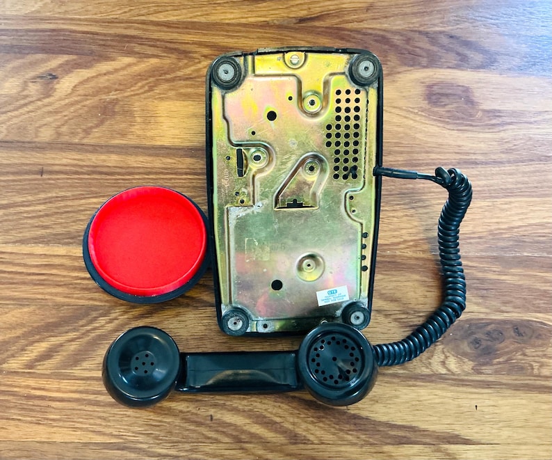 May include: A vintage black telephone with a red button and a coiled cord. The phone's base is gold-colored, and the handset is black. The phone is shown on a wooden surface.