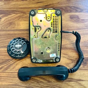 May include: A vintage black rotary dial telephone with a black handset and coiled cord. The phone's base is a metallic gold color, and the dial is black with white numbers and the word "OPERATOR". The phone is displayed on a wooden surface.