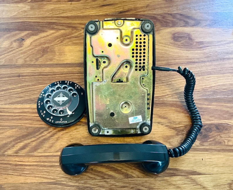 May include: A vintage black rotary dial telephone with a black handset and coiled cord. The phone's base is a metallic gold color, and the dial is black with white numbers and the word "OPERATOR". The phone is displayed on a wooden surface.