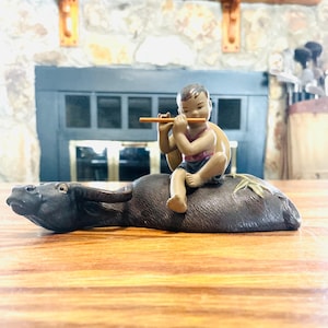 May include: A ceramic figurine depicting a young person playing a flute while riding a water buffalo. The figurine is painted with a muted color palette, featuring brown, black, and flesh tones. The water buffalo has a detailed texture, and the figure is seated on its back.