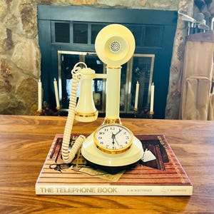 May include: Cream-colored vintage telephone with a clock face, gold trim, and a rotary dial. The phone sits on a book titled "The Telephone Book". The phone has a matching cream-colored handset and a coiled cord.