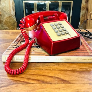May include: A glossy red landline telephone with a matching coiled cord and a gold-toned keypad featuring grey buttons. The telephone rests on a book titled The Telephone Book by H. M. Boettinger, which has a wood-grain cover design.