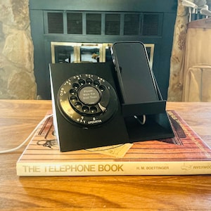 May include: A black smartphone charging dock shaped like a vintage rotary telephone base. The circular dial features the text Please Wait For Dial Tone. The dock holds a black smartphone upright and includes a white charging cable extending from the side.