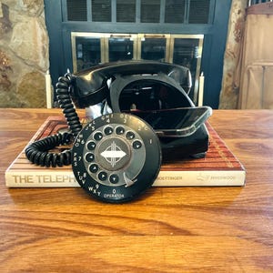 May include: A vintage black rotary dial telephone with a coiled cord, resting on a book titled "The Telephone." The dial features white numbers and the word "OPERATOR." The phone is set against a backdrop of a fireplace and wooden table.