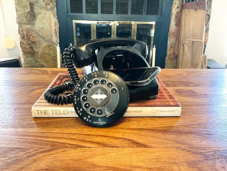 May include: A vintage black rotary dial telephone with a coiled cord, resting on a book titled "The Telephone." The dial features white numbers and the word "OPERATOR." The phone is set against a backdrop of a fireplace and wooden table.