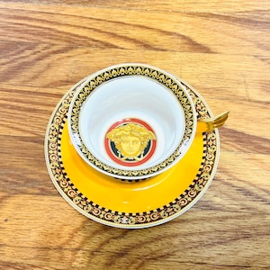 May include: A yellow teacup and saucer set with gold and black decorative trim. The teacup and saucer feature a Medusa head logo in the center. The teacup has a gold handle.