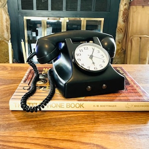 May include: A black vintage telephone with a clock face, resting on a book titled "THE PHONE BOOK" by H.M. Boettinger. The clock has white numbers and hands. The phone is on a wooden surface.