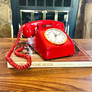 May include: A bright red vintage telephone with a clock face, resting on a book titled "PHONE BOOK". The clock has a gold-colored bezel and white face. The phone's coiled cord is red.
