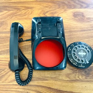 May include: A vintage black rotary phone with a red interior and a separate dial. The handset is on the left, connected to the base with a coiled cord. The dial is black with white numbers and letters.