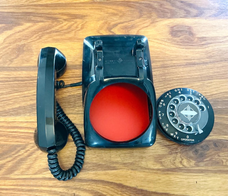 May include: A vintage black rotary phone with a red interior and a separate dial. The handset is on the left, connected to the base with a coiled cord. The dial is black with white numbers and letters.