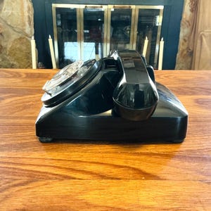 May include: A vintage black rotary dial telephone with a glossy finish. The phone features a circular dial and a curved handset. The phone is placed on a wooden surface, with a fireplace and candles in the background.