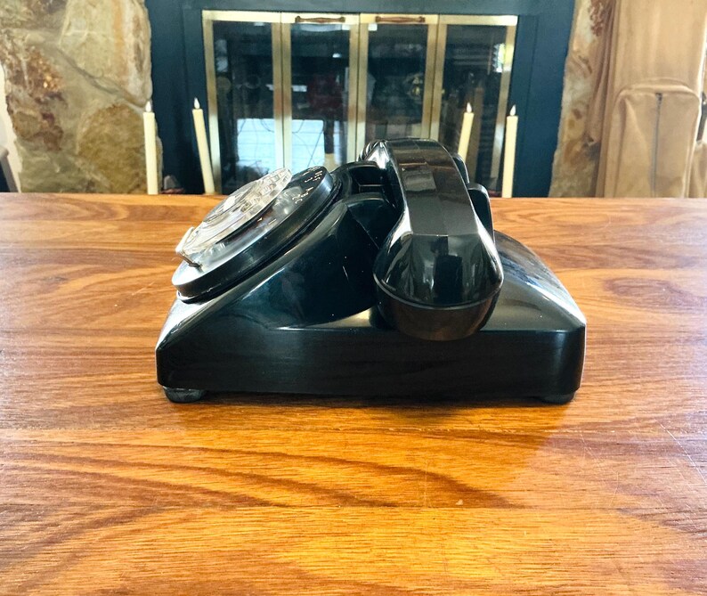 May include: A vintage black rotary dial telephone with a glossy finish. The phone features a circular dial and a curved handset. The phone is placed on a wooden surface, with a fireplace and candles in the background.