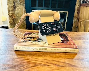 May include: A beige plastic telephone-shaped smartphone stand with a coiled cord and a handset resting on top. A black smartphone is mounted in the center, displaying a clock and a September calendar. The stand rests on a book titled The Telephone Book.