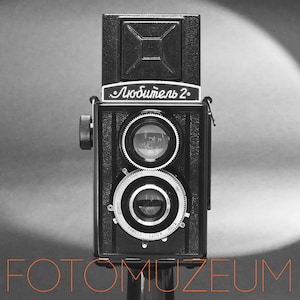 May include: A black vintage twin-lens reflex camera with the text "Любитель 2" on the front. The camera has two circular lenses and a silver dial. The word "FOTOMUZEUM" is written in orange at the bottom.