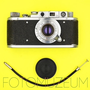 Puede incluir: Una cámara vintage en blanco y negro con lente y diales, una tapa de lente y un disparador de cable sobre un fondo amarillo. La cámara tiene el texto "ИНДУСТАР-22" y "F=50мм" en la lente. El texto "FOTOMUZEUM" está en la parte inferior.