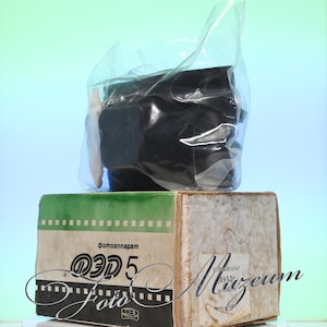 May include: A vintage black camera, sealed in a clear plastic bag, rests atop a cardboard box. The box features green and white accents, with Cyrillic text and the word "Foto" in a decorative font. The overall aesthetic suggests a retro or antique item.