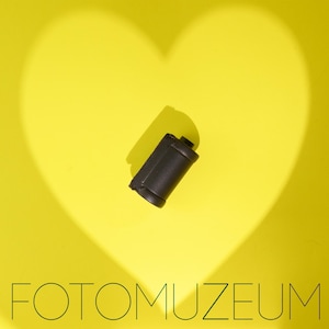 Puede incluir: Un carrete de película negro de 35 mm sobre un fondo amarillo brillante con una sombra en forma de corazón. La palabra "FOTOMUZEUM" está impresa en negro en la parte inferior de la imagen.