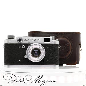 May include: A vintage ФЭД-2 camera with a black leather body and silver accents. The camera has a lens with measurement markings and is accompanied by a brown leather case. The camera is displayed on a white surface with the text "Foto Muzem".