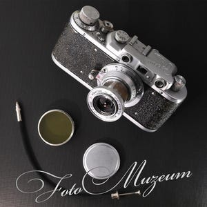 Puede incluir: Una cámara vintage plateada y negra con lente, filtro y tapa. La cámara tiene varios diales y un agarre texturizado. También hay un cable negro y una pequeña herramienta. El texto "Foto Muzeum" es visible.