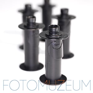 May include: Several black film canisters are arranged against a white background. Each canister has a circular base and a threaded top. The text "FOTOMUZEUM" is displayed in blue and orange at the bottom of the image.