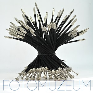 May include: A bundle of black pen refills with silver-colored metal tips. The refills are tied together with a black rubber band. The image has the text "FOTOMUZEUM" in blue at the bottom.