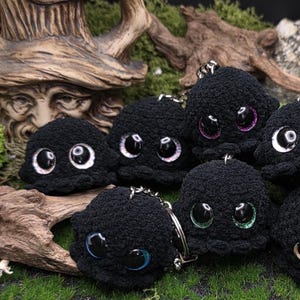 Könnte beinhalten: Eine Sammlung schwarzer gehäkelter Oktopus-Schlüsselanhänger mit großen, ausdrucksstarken Augen in verschiedenen Farben, darunter Weiß, Rosa, Blau und Gold. Die Schlüsselanhänger werden auf grünem Moos und Holz präsentiert.