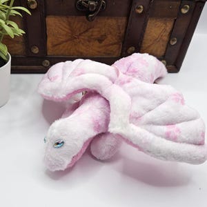 Könnte beinhalten: Ein rosa-weißes Drachen-Plüschtier mit Blumenmustern. Der Drache hat große, weiche Flügel und einen langen Hals. Die Augen sind blau und das Spielzeug besteht aus weichem, flauschigem Material. Der Drache liegt auf einer weißen Oberfläche.