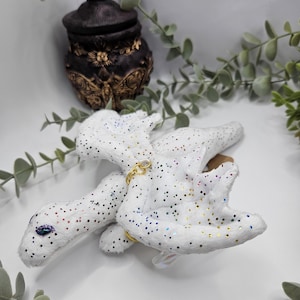 Könnte beinhalten: Ein weißes Plüschdrachenspielzeug mit schillernden Glitzerpunkten und einer goldfarbenen Kette. Der Drache hat ein blaues Auge und liegt auf einer weißen Oberfläche. Im Hintergrund befindet sich ein dekoratives braunes Glas mit goldenen Akzenten.